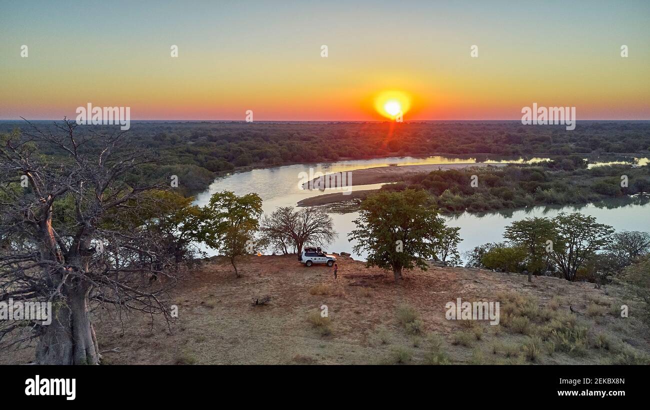 Aerial view of a jeep with a rooftop tent close to the river and a big baobab tree at sunset, Cunene river area, Angola Stock Photo