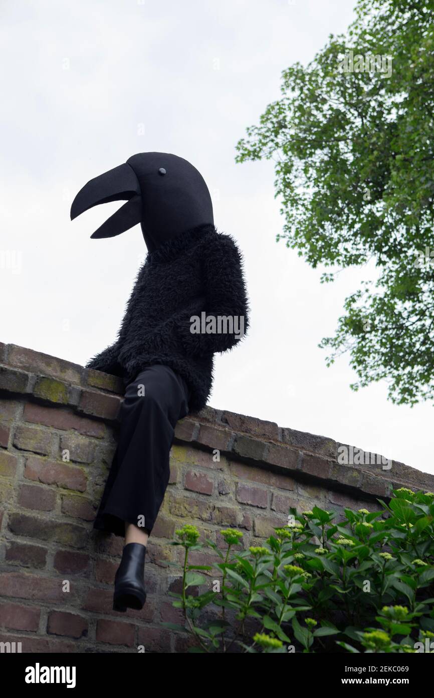Female wearing crow costume sitting on retaining wall by tree Stock Photo