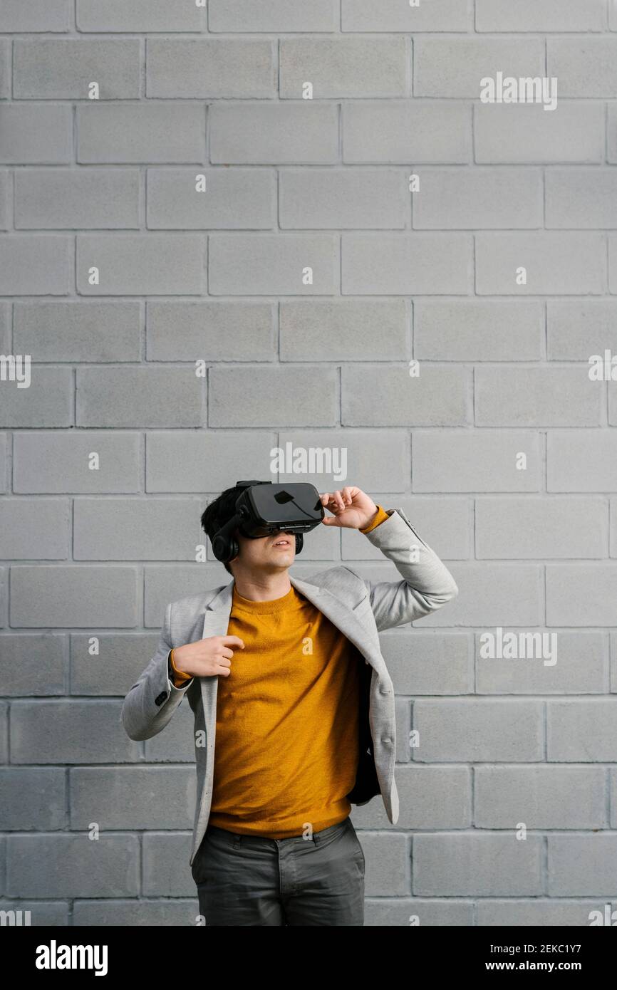 Young man wearing blazer using virtual reality headset while standing against gray color Stock Photo