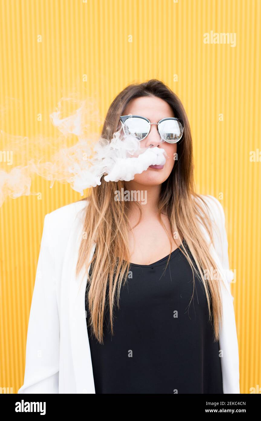 Young woman exhaling smoke against yellow wall Stock Photo