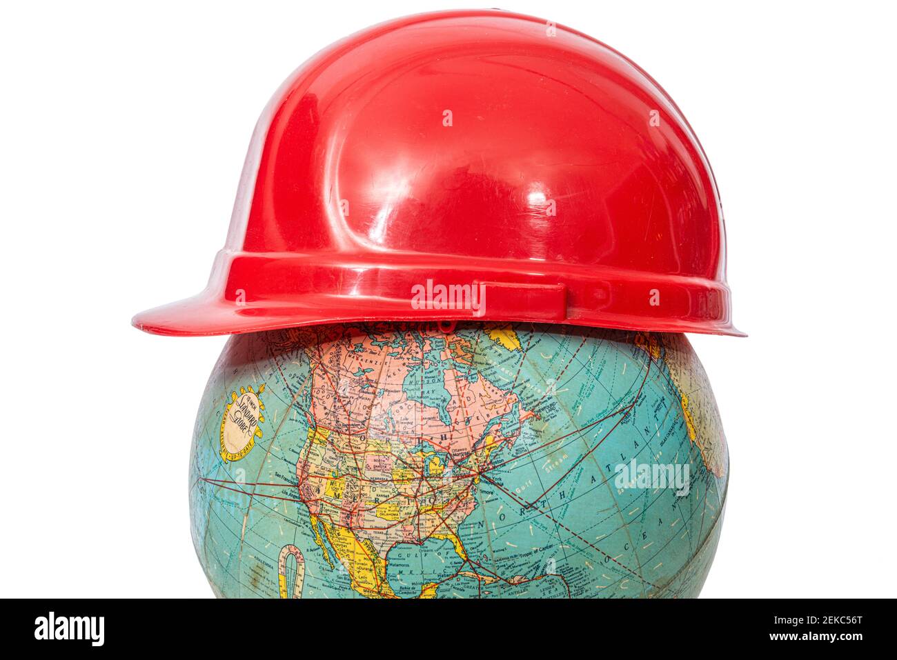 Horizontal shot of a side view of an old red hard hat sitting on top of a pre world war 2 world globe.  White background. Stock Photo