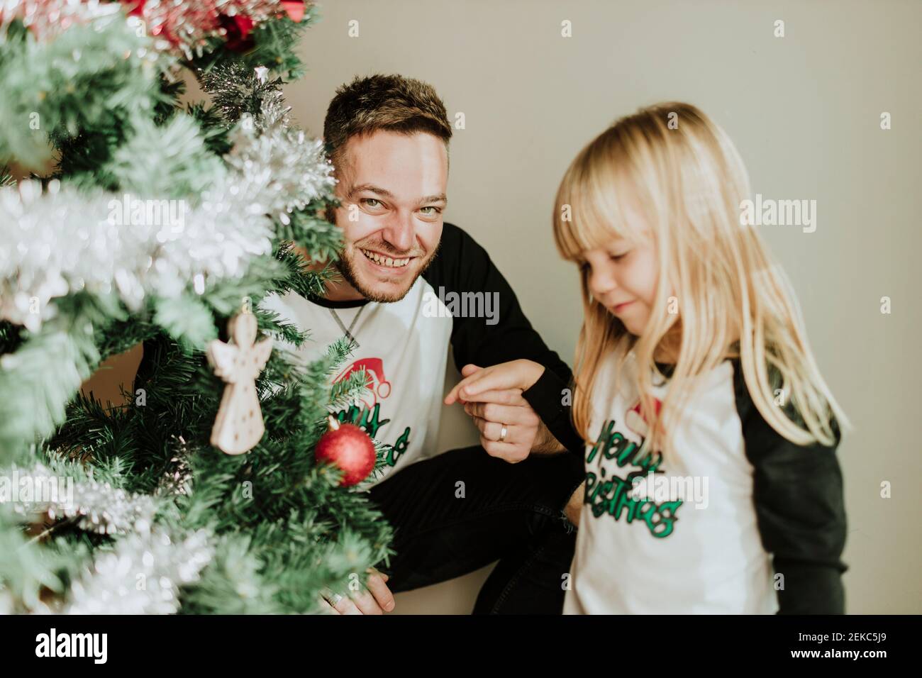 Girl showing Christmas ornament at camera while family decorating Christmas tree in home Stock Photo