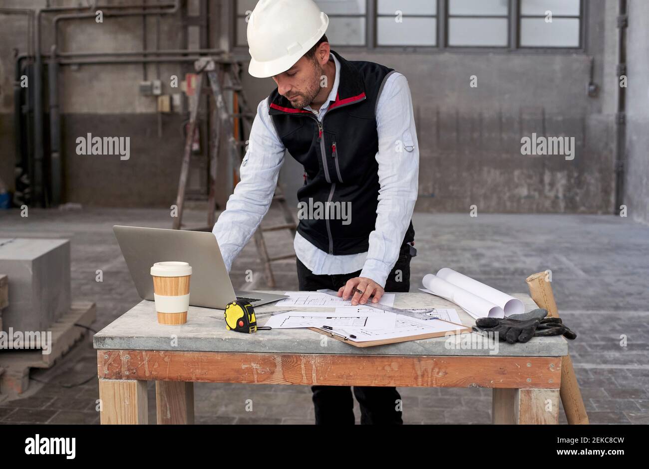 Male architect with blueprints using laptop on table while standing in building Stock Photo