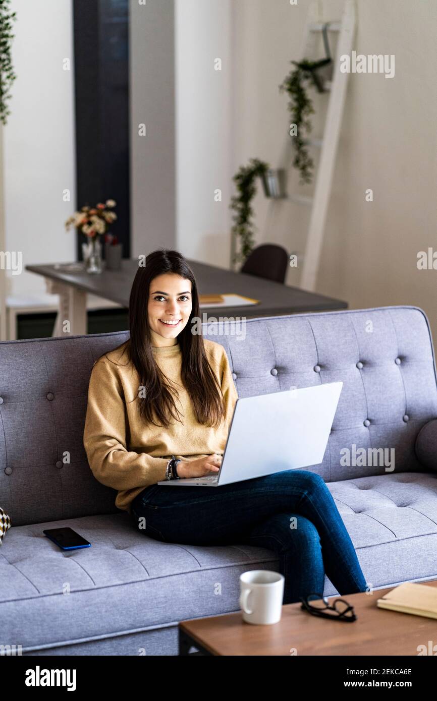 Smiling woman using laptop while sitting with legs crossed at knee on sofa at home Stock Photo