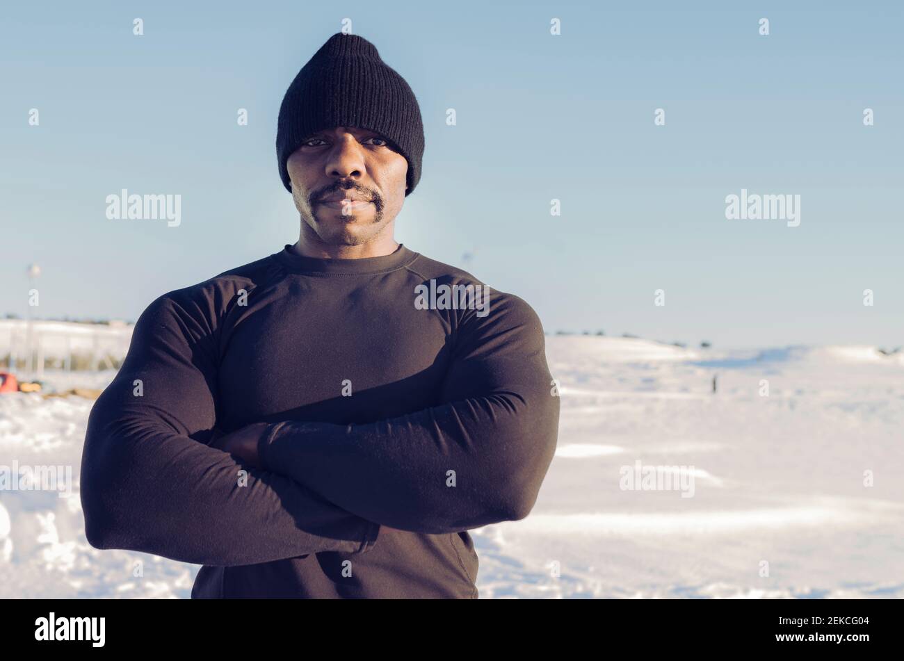Muscular build sportsman wearing knit hat standing with arms crossed in snow during sunny day Stock Photo
