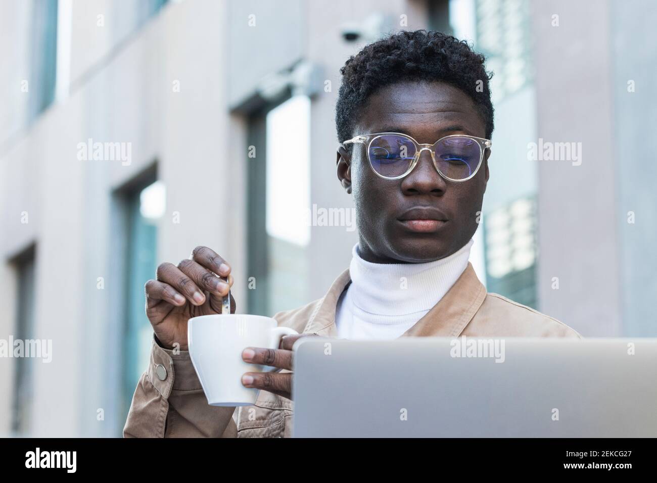 Young man stirring coffee while looking at laptop at sidewalk cafe Stock Photo