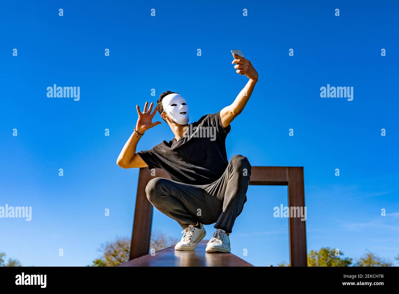 Young man wearing white mask waving hand to video call through mobile phone while crouching on retaining wall Stock Photo