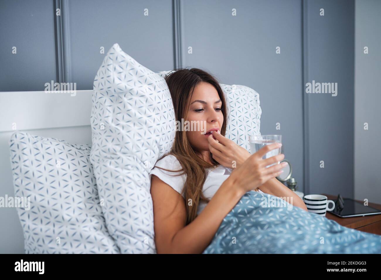 Close up of cute stressed young girl taking medications with water while lying ill in the bed at home. Stock Photo