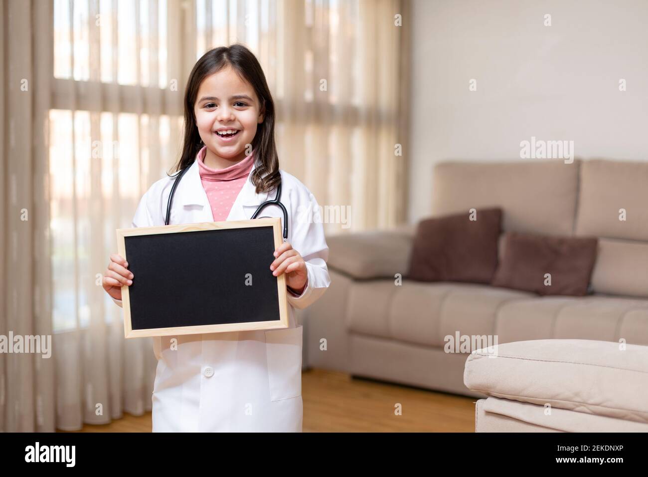 Smiling little child in doctor's uniform holding a blackboard at home. Space for text. Stock Photo