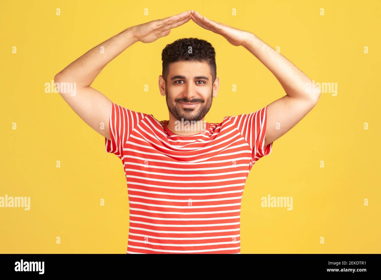 I'm in safety. Portrait of happy bearded man in striped t-shirt standing raising hands showing roof gesture and smiling contentedly, dreaming of house Stock Photo