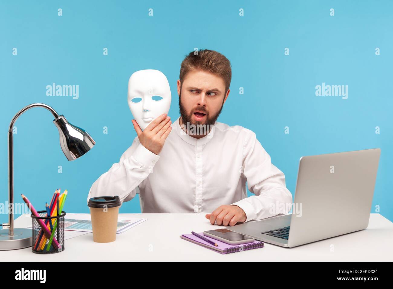 Bearded man office worker looking at white face mask in his hand with suspicion, hiding his identity, spying undercover, corruption. Indoor studio sho Stock Photo