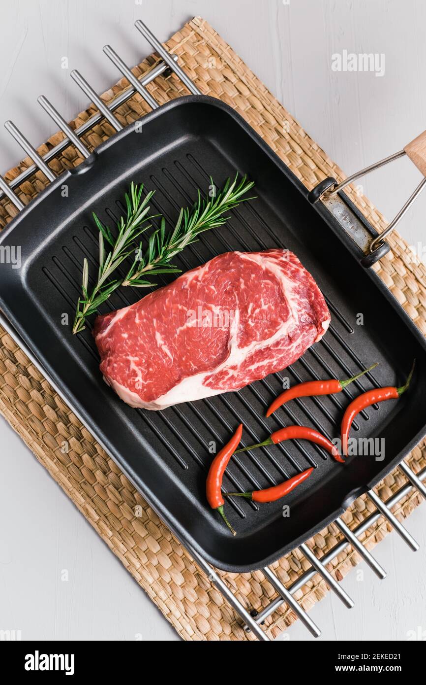 Raw rib eye beef steak on black grilling pan with chili pepper and rosemary, top view. Stock Photo