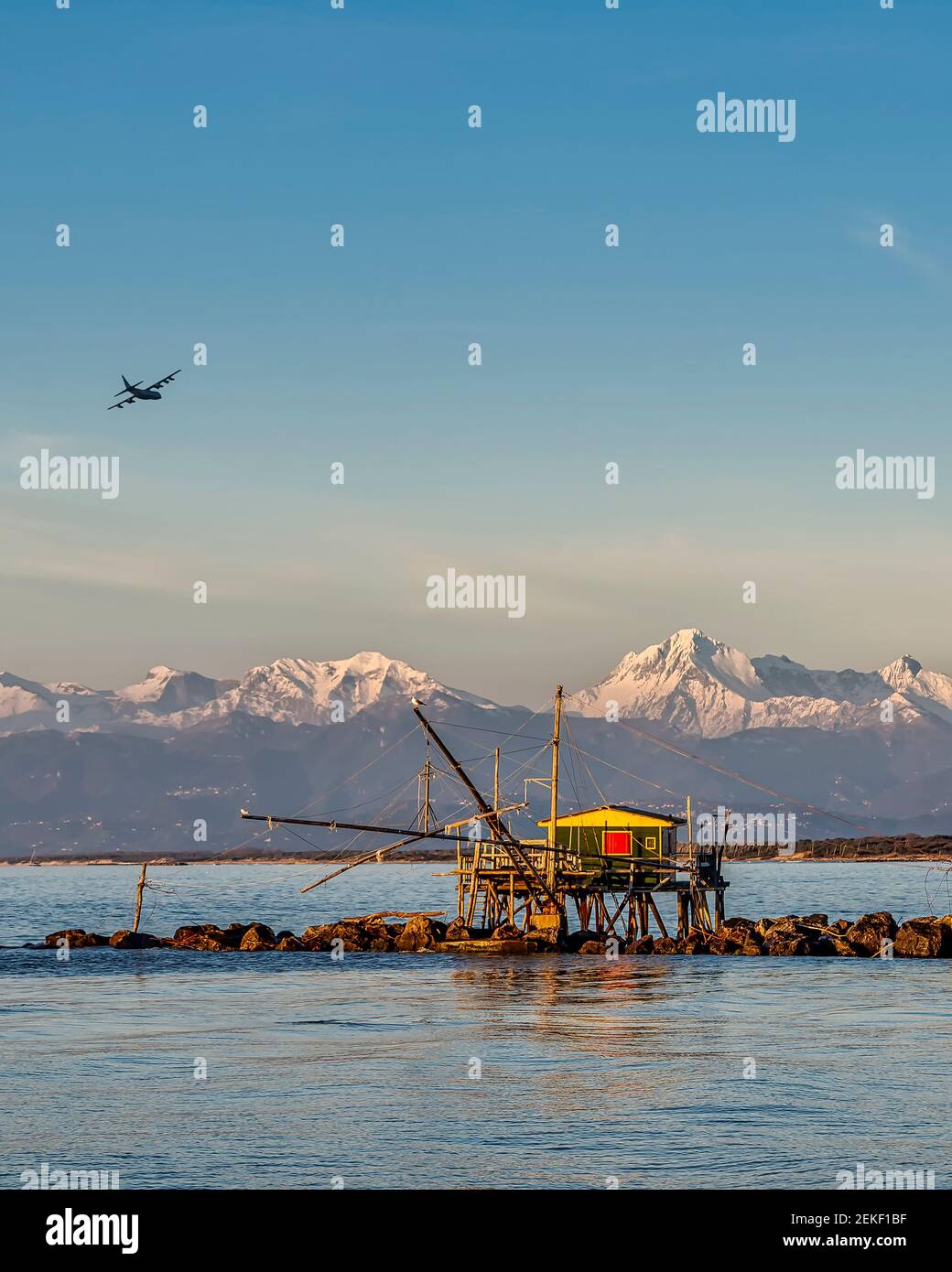 An airplane flies over a trebuchet fishing hut at sunset, against the snow-capped Alps, Marina di Pisa, Tuscany, Italy Stock Photo