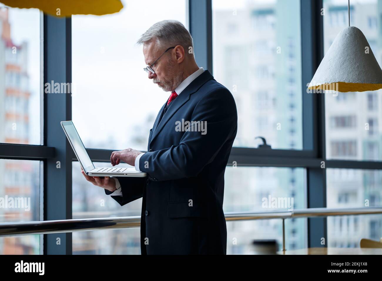 Smiling happy managing director thinks about his successful career development while standing with a laptop in his office near the background of a win Stock Photo