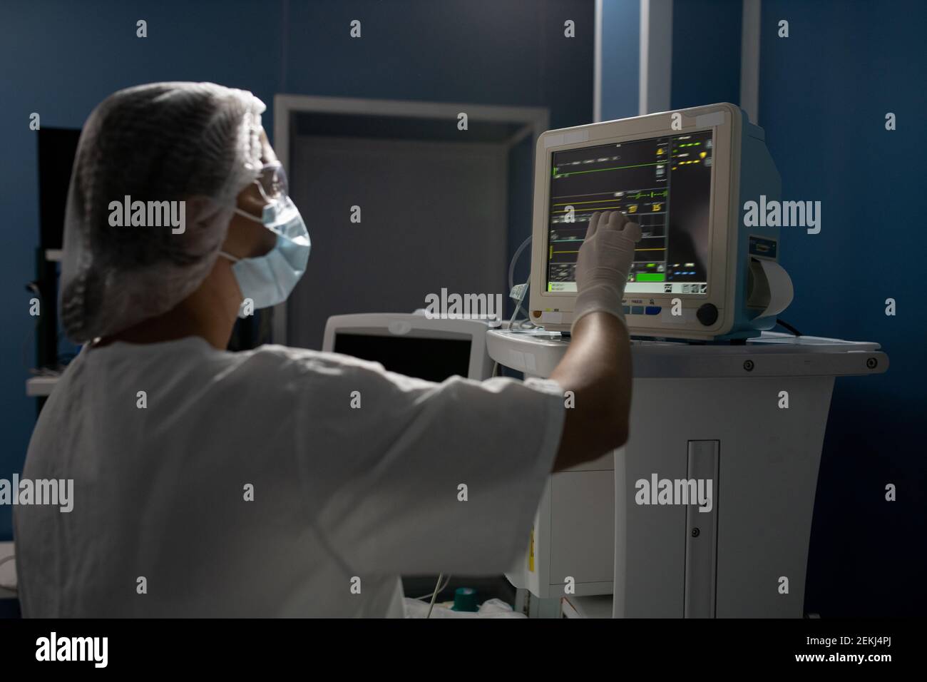 Young female assistant in gloves, mask and uniform pointing at display of medical equipment while taking control of patient body condition Stock Photo