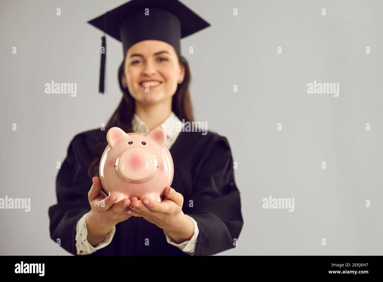 Pink piggy bank in the hands of a joyful female student standing on a gray background. Stock Photo