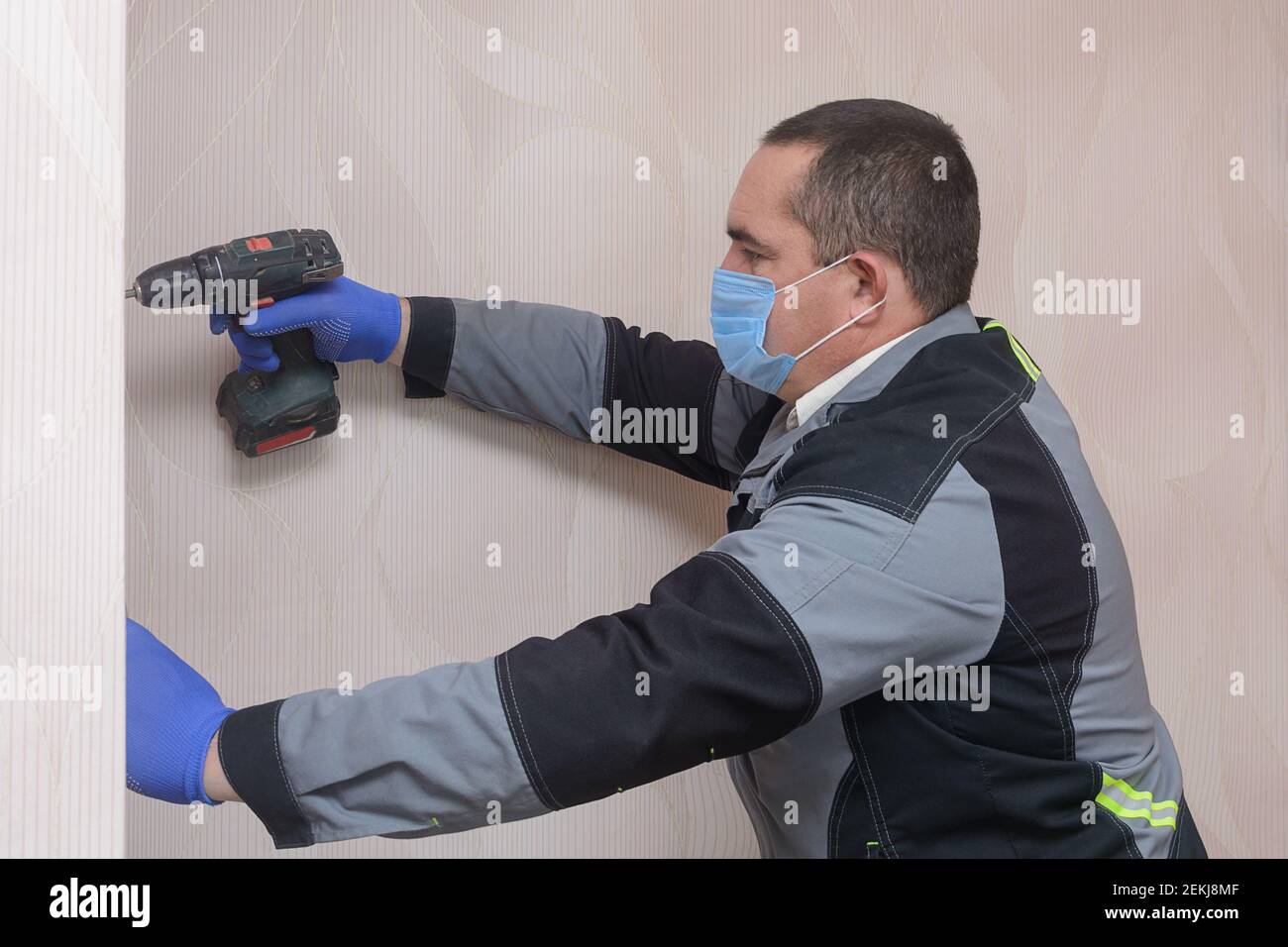 Repairman in overalls works as screwdriver. Master wearing  medical mask and gloves complies with safety rules during pandemic. Stock Photo