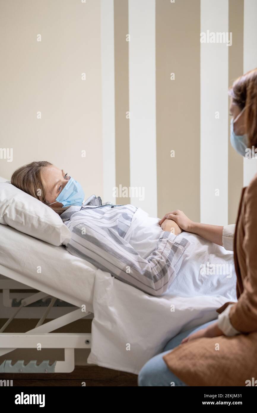 Young sick female patient in pajamas and protective mask lying in bed in hospital chamber and looking at her friend during conversation Stock Photo