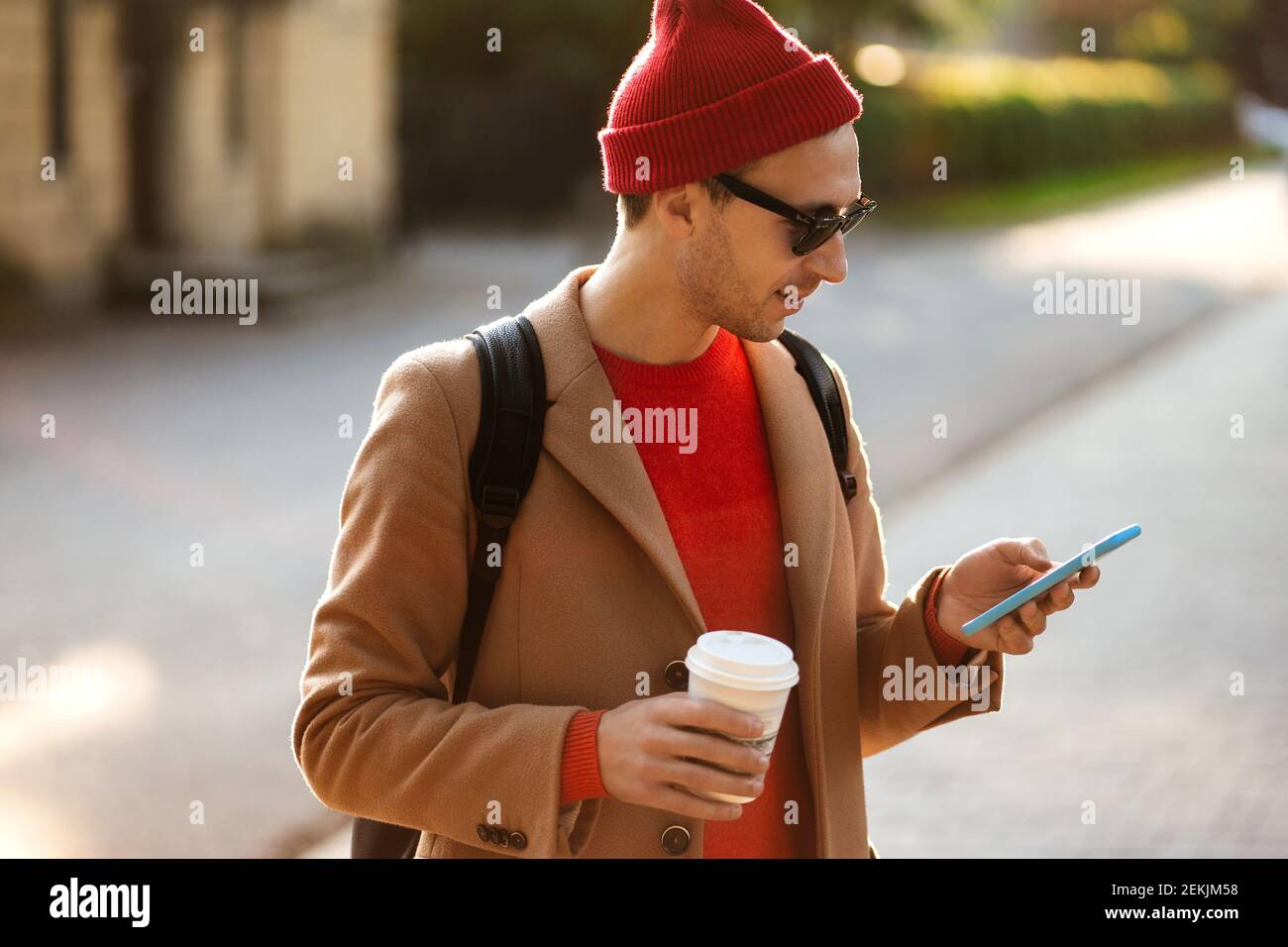 Smiling hipster man wear sunglasses using cellphone standing at the street drinking takeaway coffee  Stock Photo