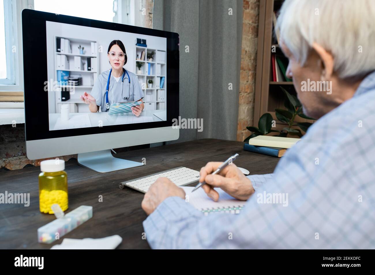 Sick senior man writing down advice of doctor while sitting in front of computer monitor where young woman on screen consulting him Stock Photo