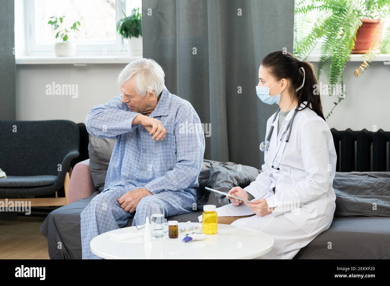 Young female general practitioner with touchpad looking at sick senior man going to cough or sneeze while sitting on couch in front of her Stock Photo