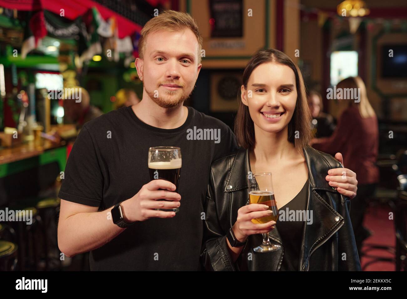 Portrait of young couple holding glasses of beer and smiling at camera while standing in the bar Stock Photo