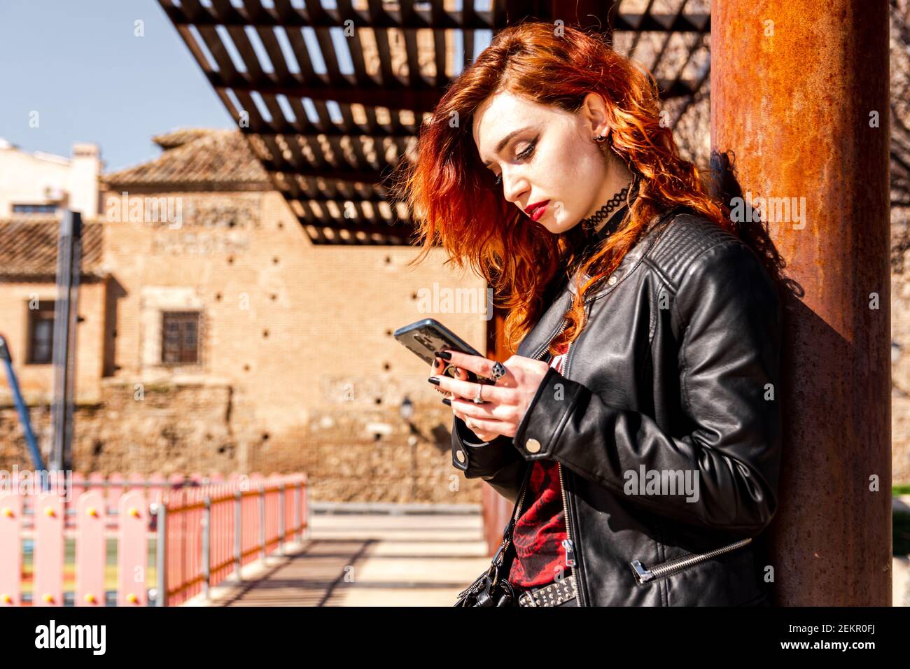 Alternative girl with blue eyes and orange hair leaning on a column while using her mobile phone. Stock Photo