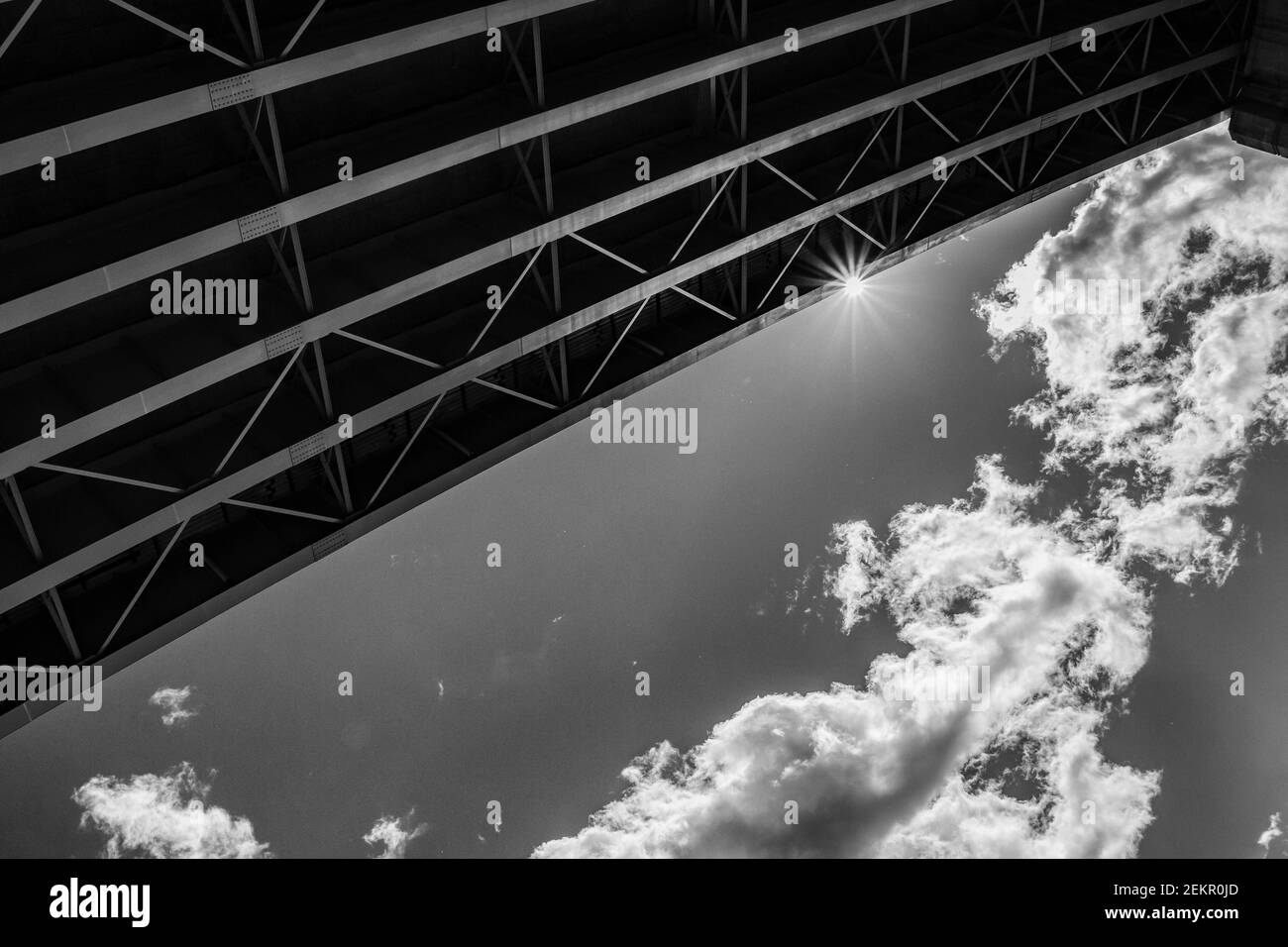 The lines of steel beams underneath the overpass create a striking contrast to the sky, clouds, and sun flare. Stock Photo