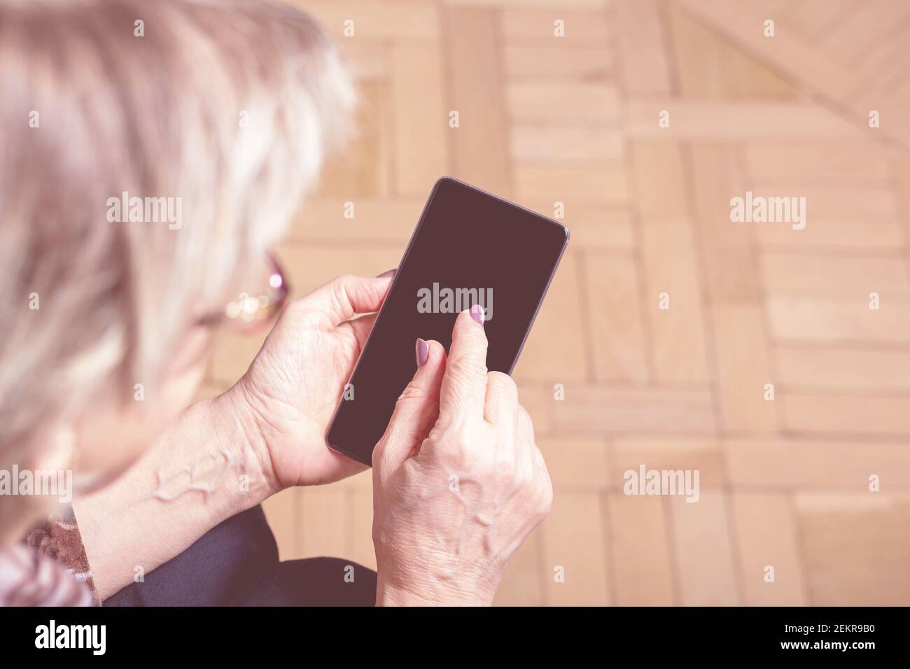 Elderly womans hand holding mobile cell phone. Over shoulder. Old woman looking at cell phone. Close-up. Blanck mobile phone screen. Display media.  Stock Photo