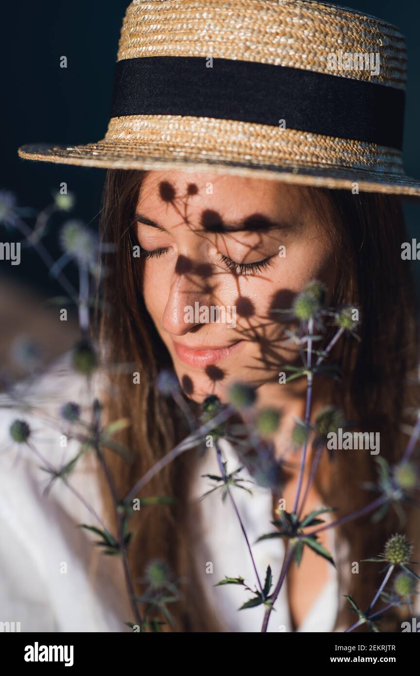 Young tender brunette girl wearing hat with a herbal branch in front of her face. Portrait of a woman with a flower, artistic shadow drops on her face Stock Photo