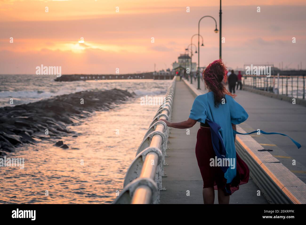 Woman in blue dress and with pink hair at sunset on St Kilda beach pier