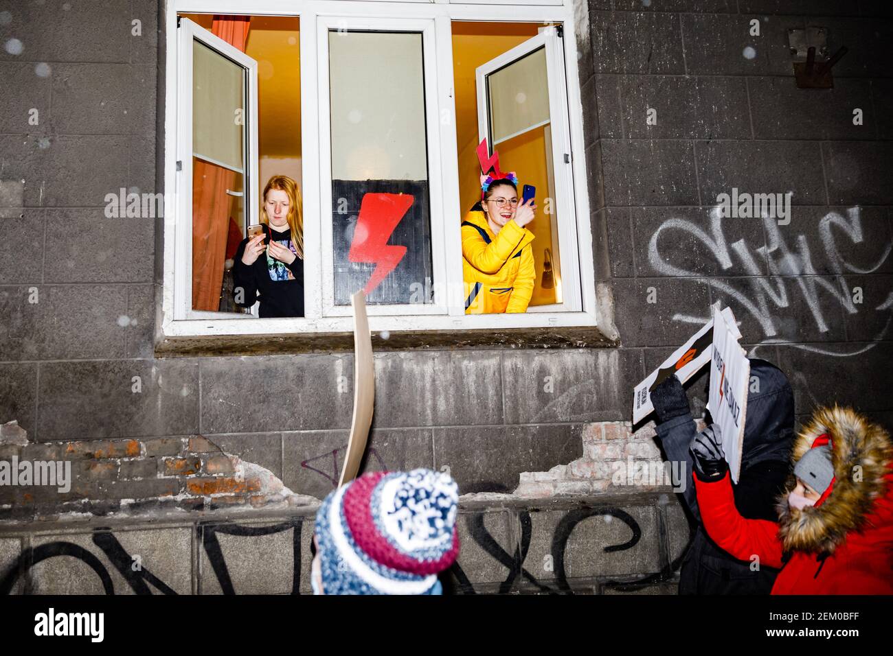 Two young women are seen with smarphones taking photos of the protesters through the opened windows of their flat. A red lighting, the symbol of Women Stock Photo