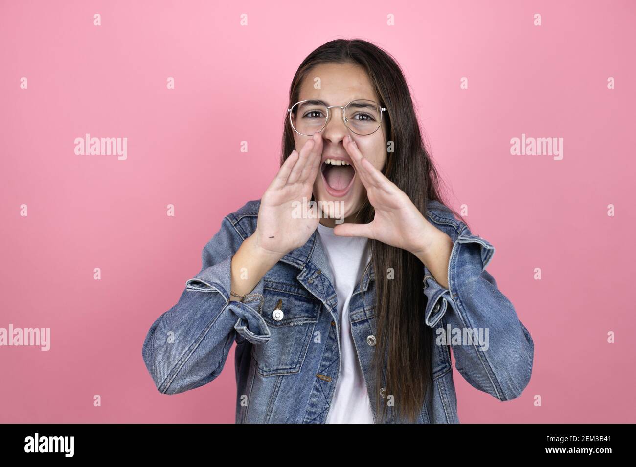 Beautiful child girl with long hair wearing a denim jacket standing over isolated pink background shouting and screaming loud to side with hands on mo Stock Photo