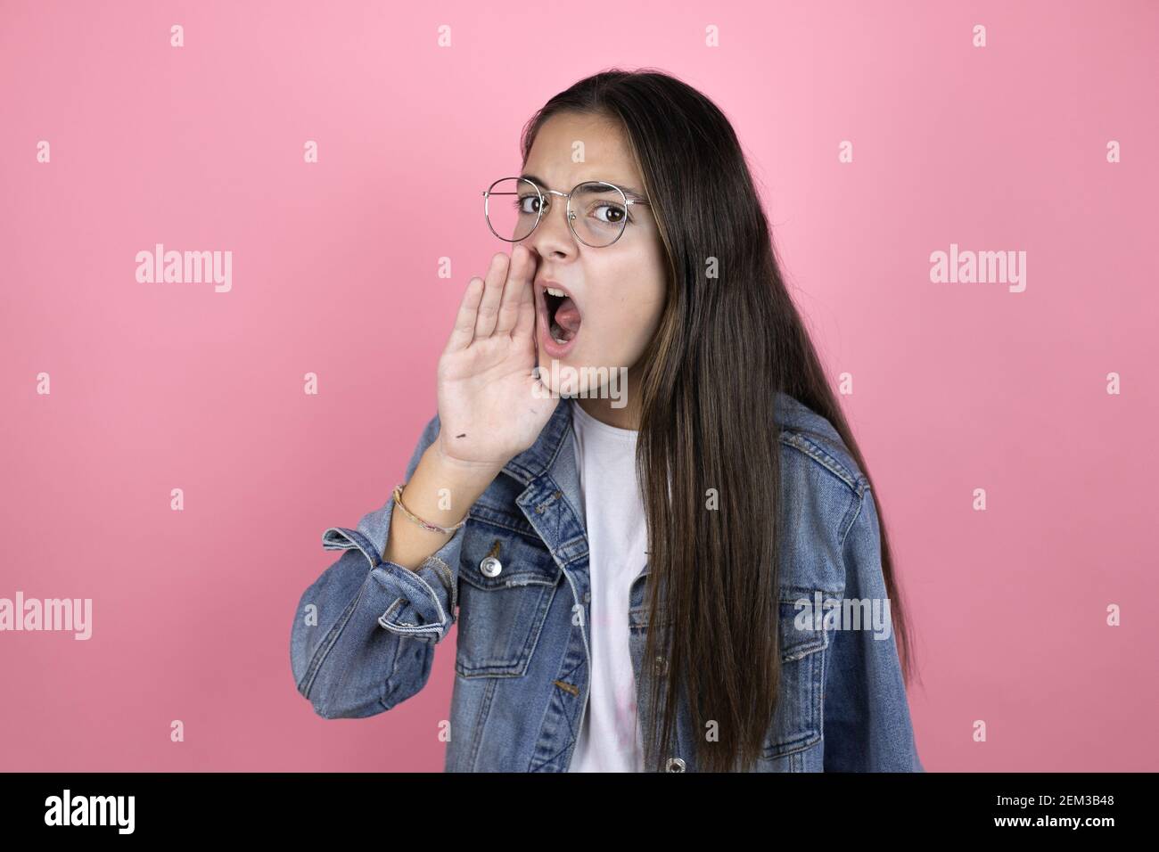 Beautiful child girl with long hair wearing a denim jacket standing over isolated pink background shouting and screaming loud to side with hand on mou Stock Photo