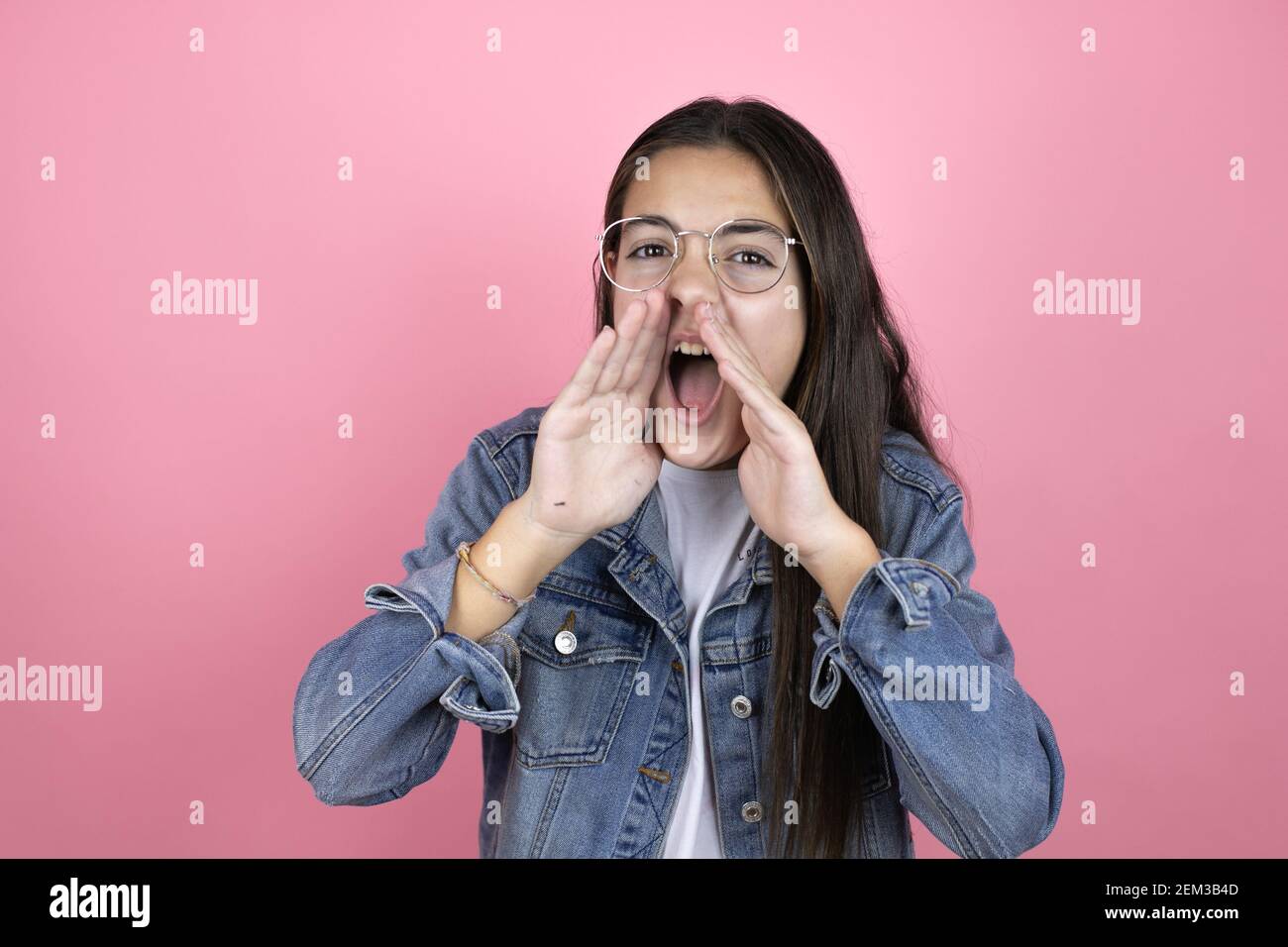 Beautiful child girl with long hair wearing a denim jacket standing over isolated pink background shouting and screaming loud to side with hands on mo Stock Photo