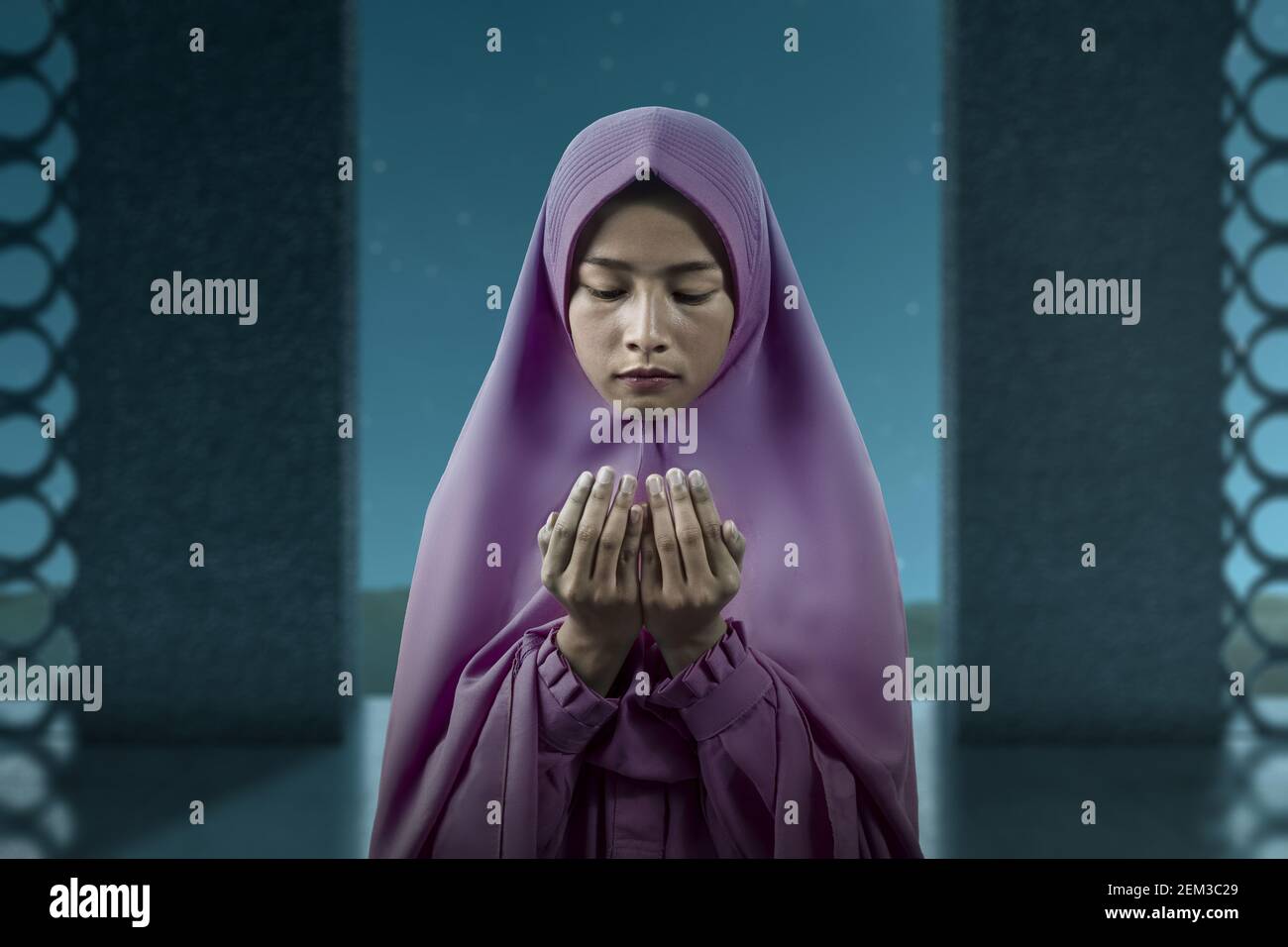 Asian Muslim woman in a veil standing while raised hands and praying on ...