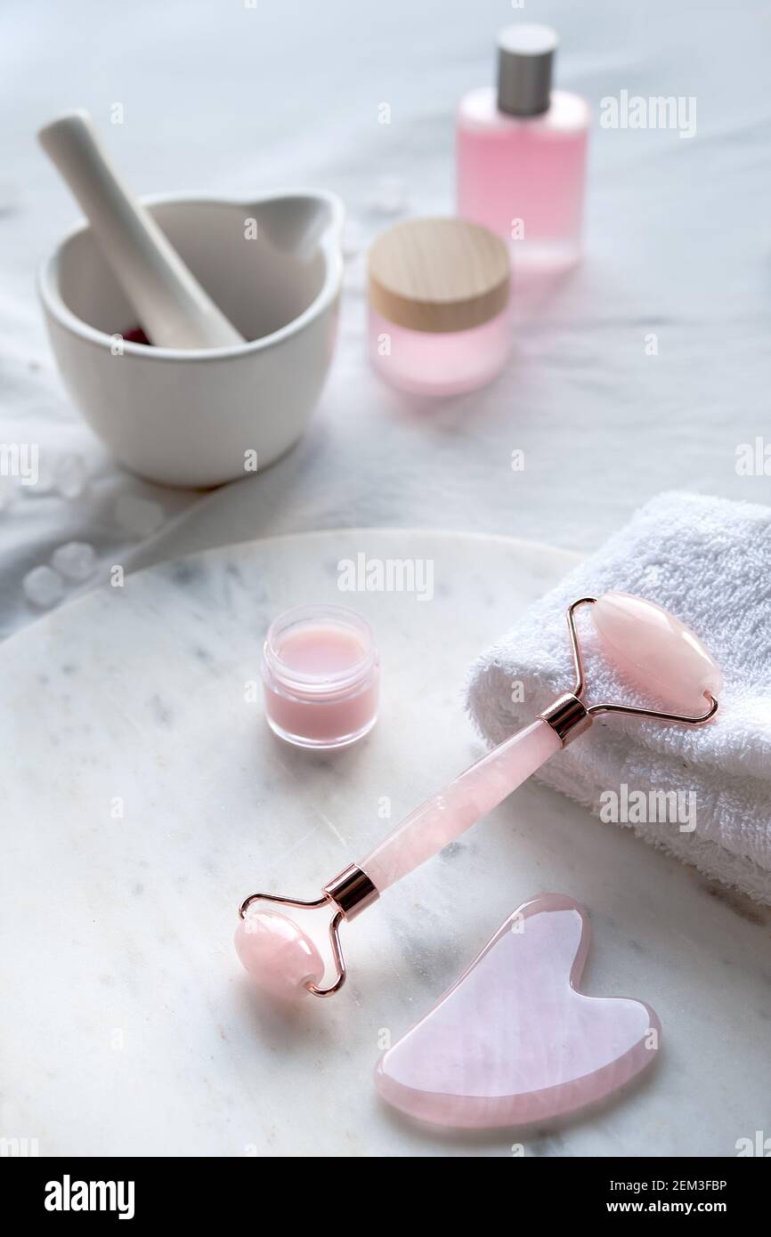 Crystal rose quartz facial roller, Gua sha stone for face massage. Pink skin care products and oils. Off white marble and textile background, natural Stock Photo
