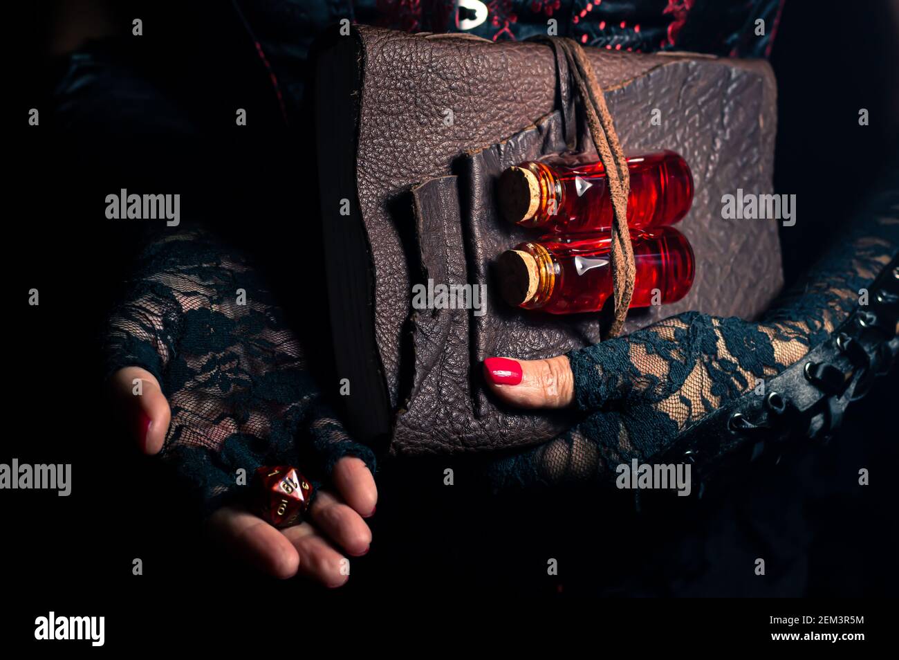 Women wearing black lace gloves holding a brown leather bound tome with glass bottles containing red liquid and a 20 sided die Stock Photo