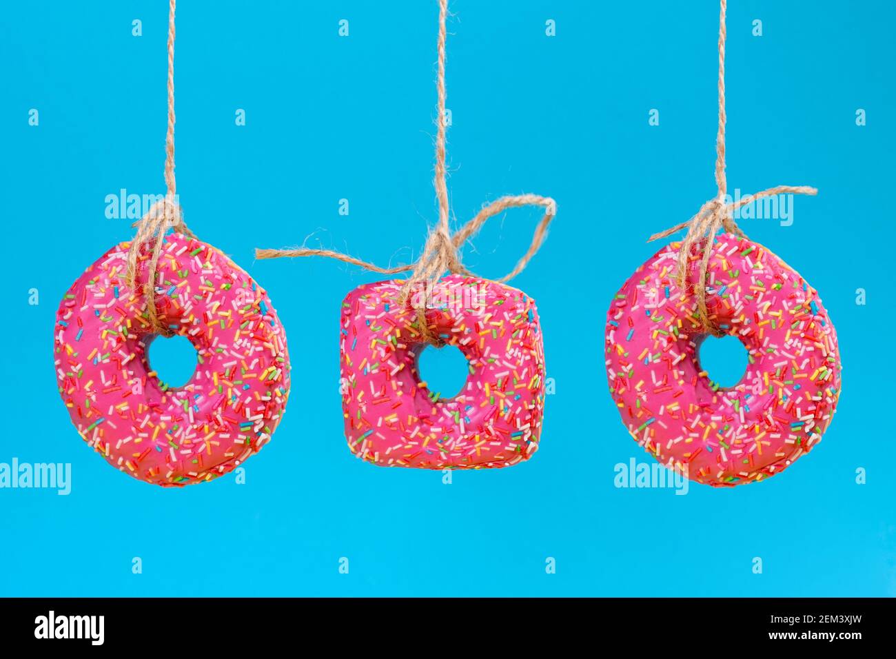 Three doughnuts of different shapes with pink frosting, suspended on a string. Close-up. Blue background. Stock Photo