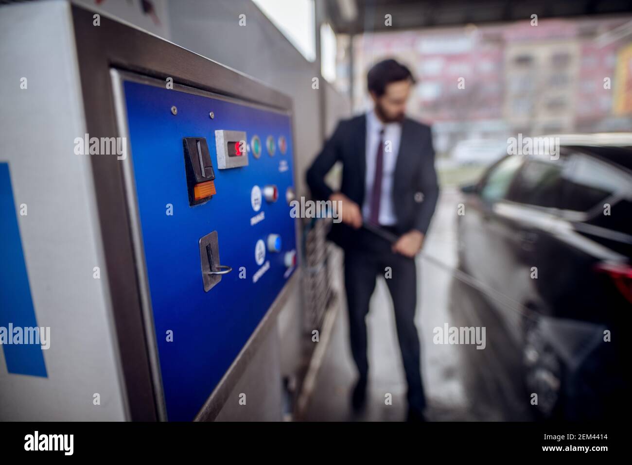 Close up focus view of coin machine while stylish bearded young hardworking man in suit washing his black car with a water gun on the manual self-serv Stock Photo