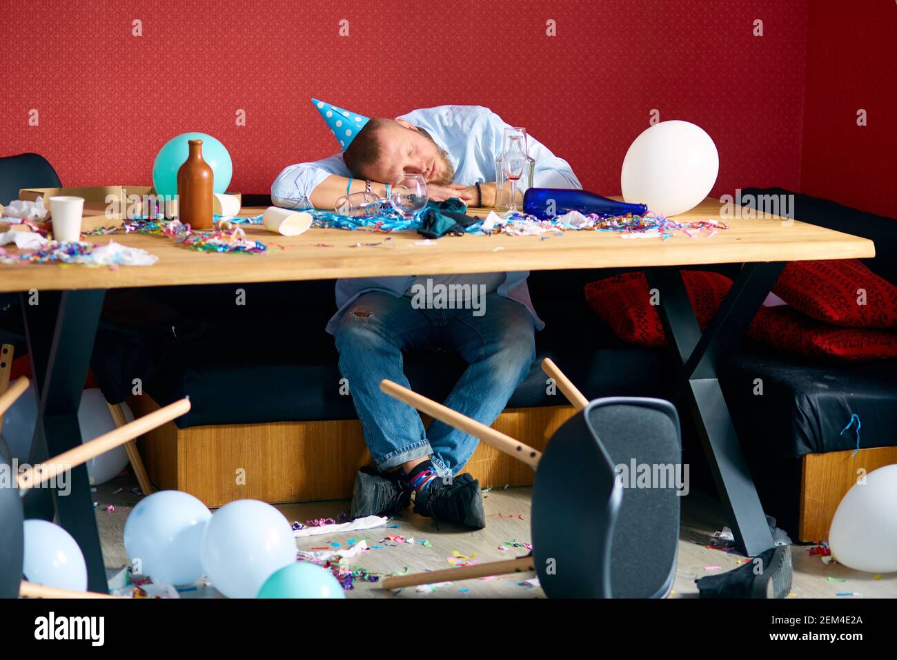 Man sleeping at table with blue cap in messy room after birthday party
