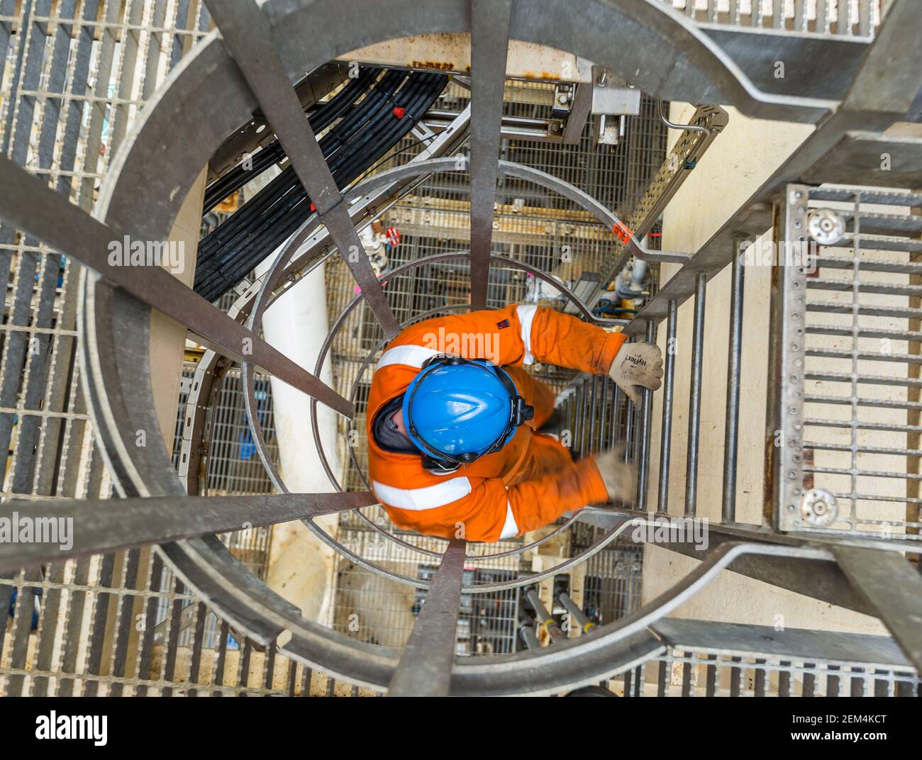 A worker wearing a boiler suit, hard hat, and gloves descending a vertical ladder with safety cage Stock Photo