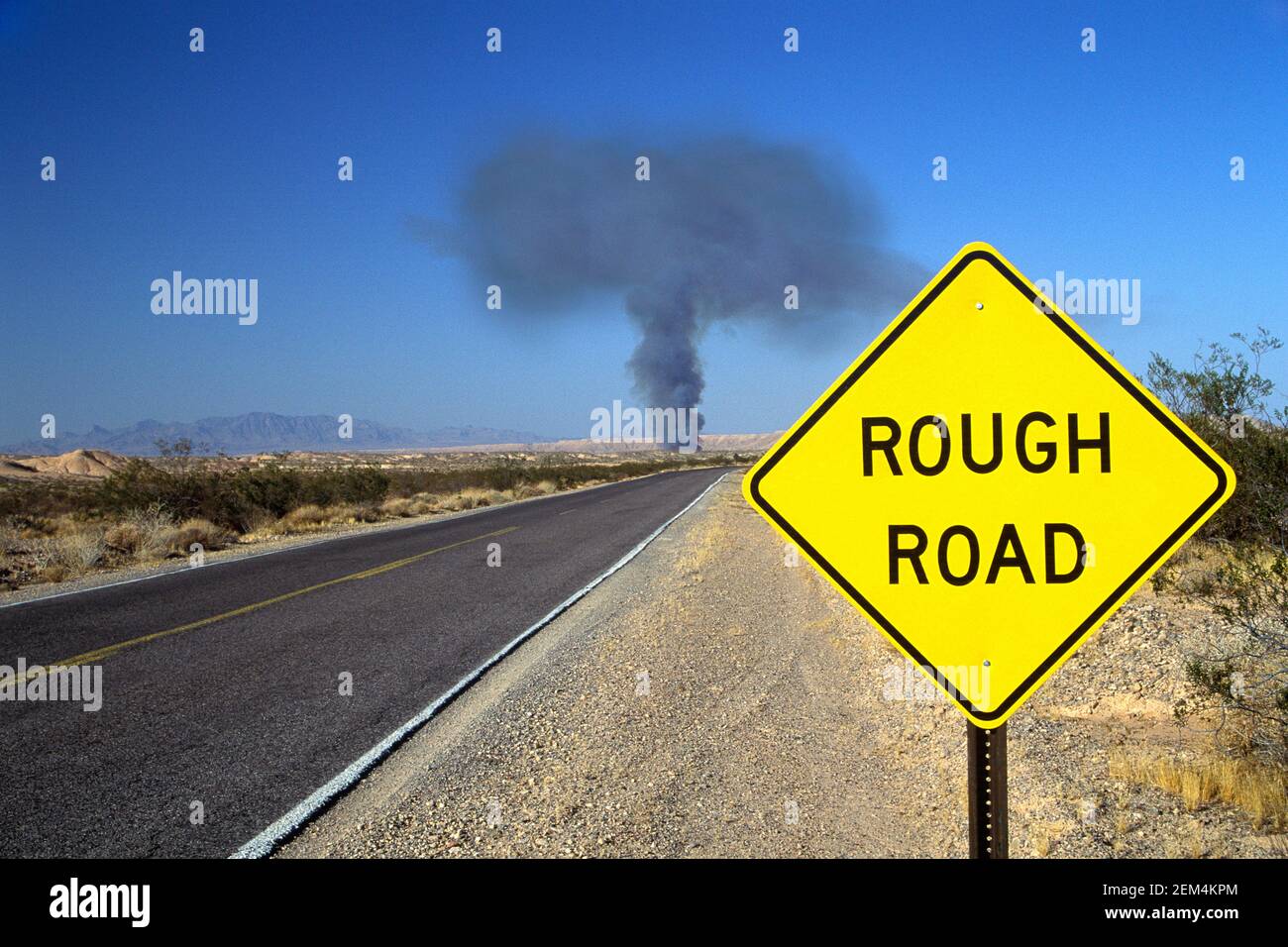 'Rough Road' ahead, yellow highway sign in front of mushroom shaped smoke cloud on road in western US. Concept: 'trouble ahead' Stock Photo