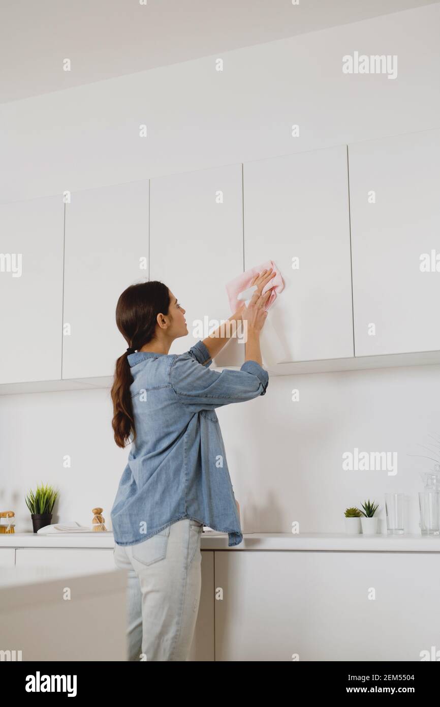 Back view of young woman cleaning a surface of white kitchen wall cabinet with rag and spray bottle detergent. Spring cleaning concept. Stock Photo