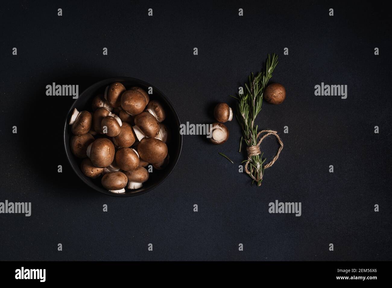 Bowl of raw baby bella mushrooms and bunch of fresh rosemary on dark grey background viewed from