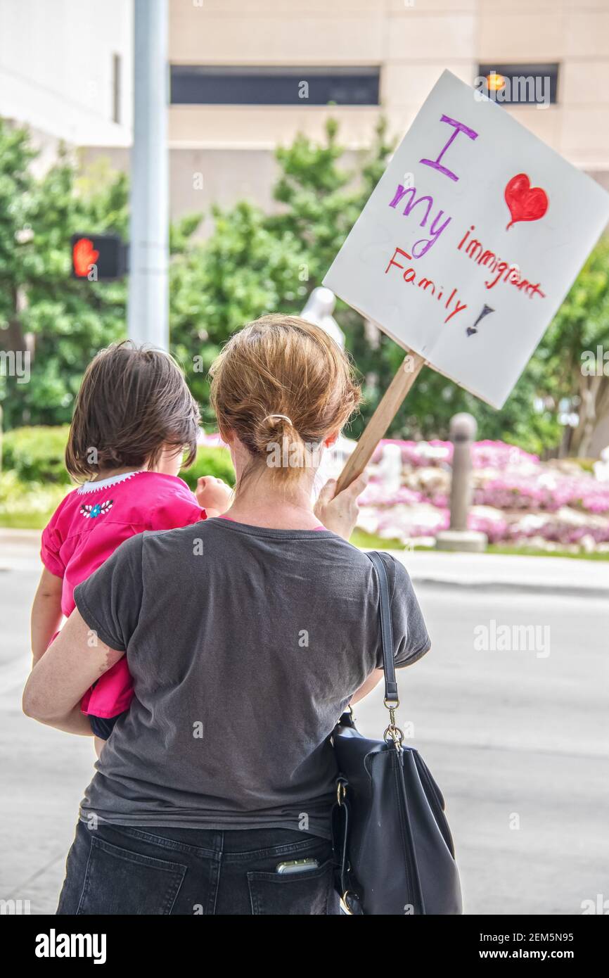 Woman with blond ponytail in black jeans  with phone in back pocket and tee-shirt holds little daughter and a sign saying I love my immigrant family o Stock Photo
