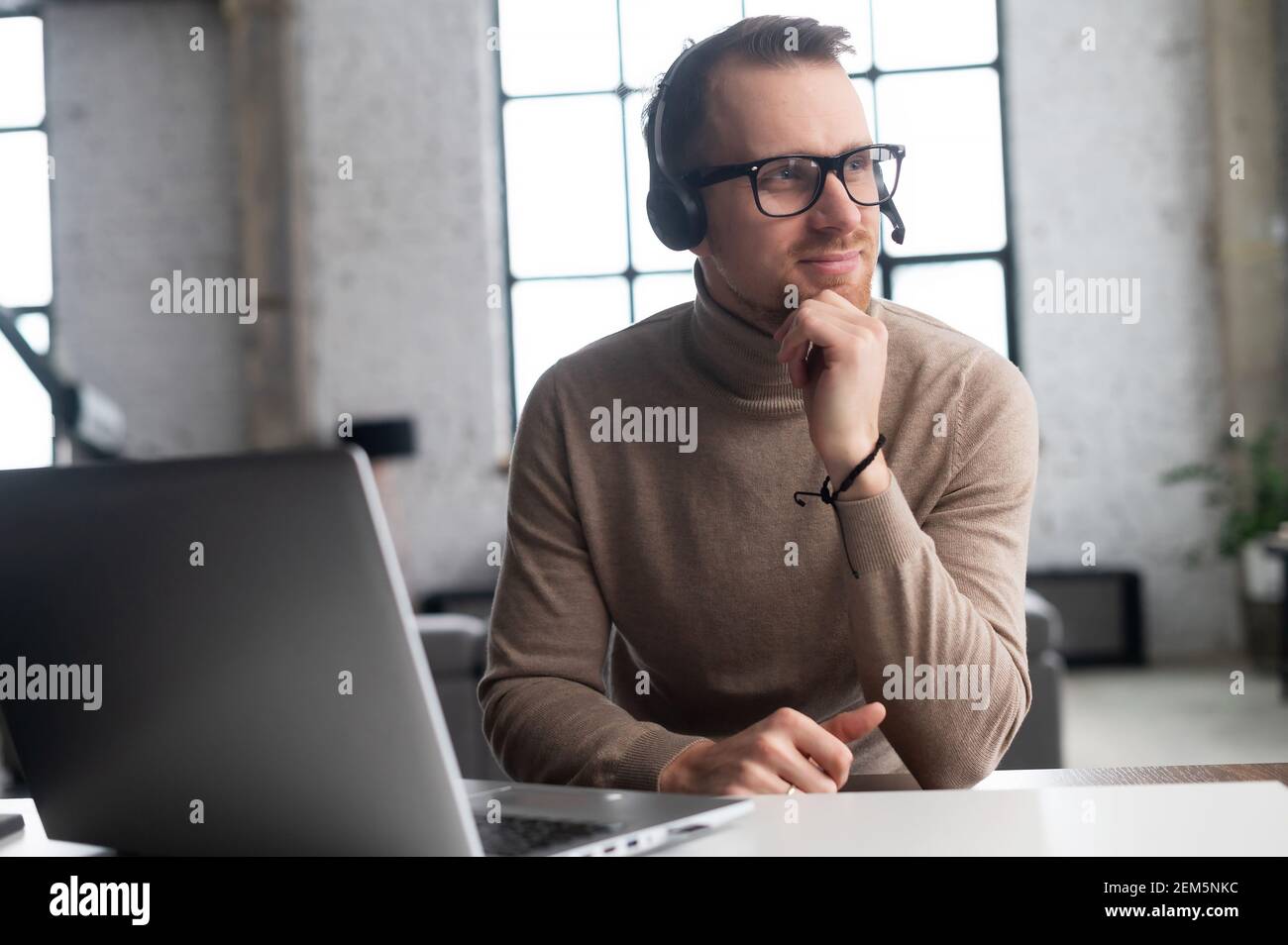 Confident worker with headset on the break, at the laptop, looking away, thinking about his family, nice memory with smile, rests his chin with the hand, in modern office, dressed in beige sweater Stock Photo