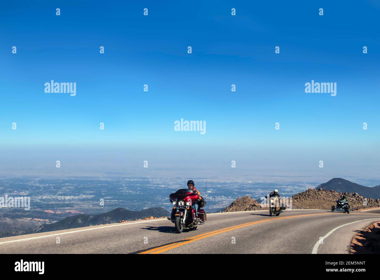 Three helmeted motorcycle drivers in a row round a curve above the tree line on the road up Pikes Peak Colorado USA with a panorama spread out way bel Stock Photo