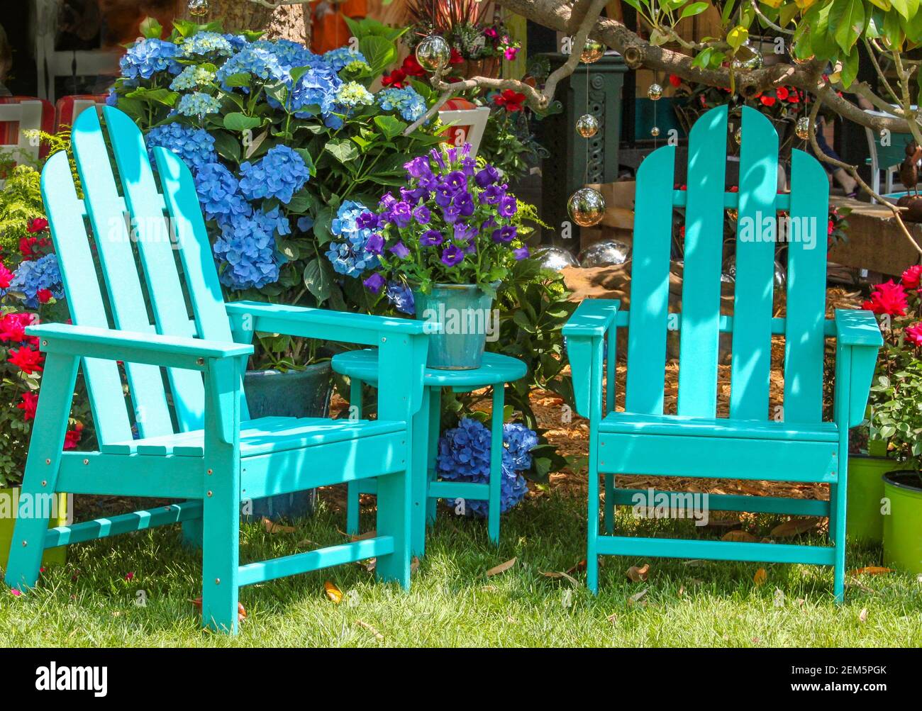 Two turquoise Adirondack chairs and a matching table surrounded by beautiful flowers and trees and shining mirror balls handing from the branches abov Stock Photo