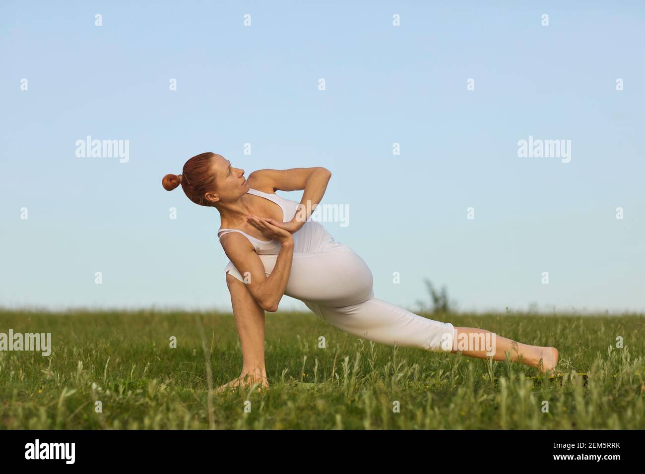 Slim female doing Revolved Crescent Lunge while practising yoga on green summer meadow Stock Photo
